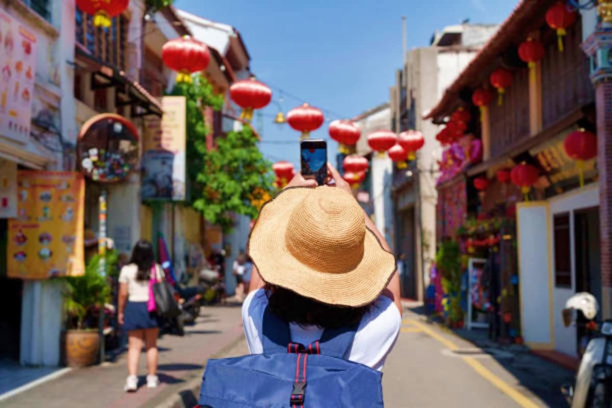 Tourist wearing straw hat photographing red lanterns hanging above a historic Chinese street during daytime in Malaysia