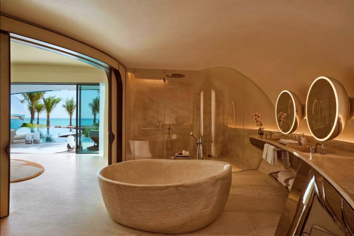 Luxury bathroom with oval stone bathtub, dual illuminated mirrors, and ocean view through glass doors overlooking palm trees 