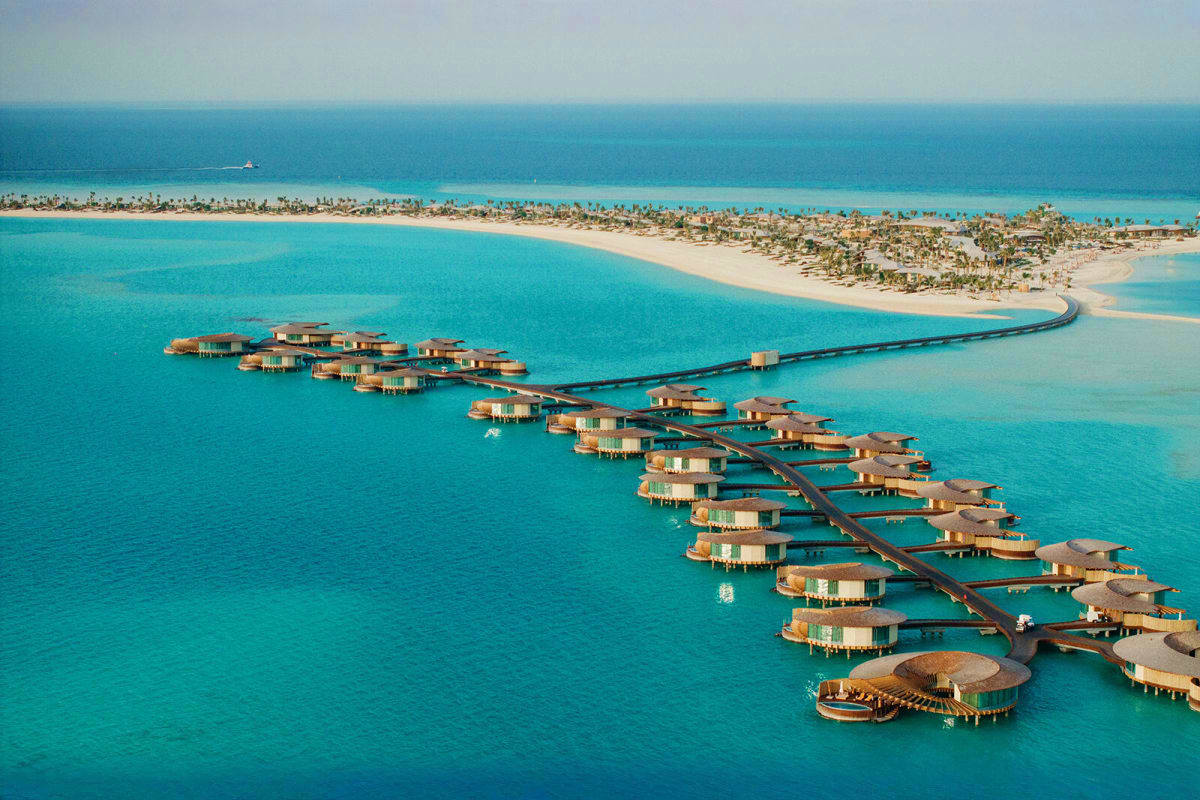 Aerial view of overwater bungalows connected by wooden boardwalks in turquoise lagoon waters with sandy beach and palm trees 