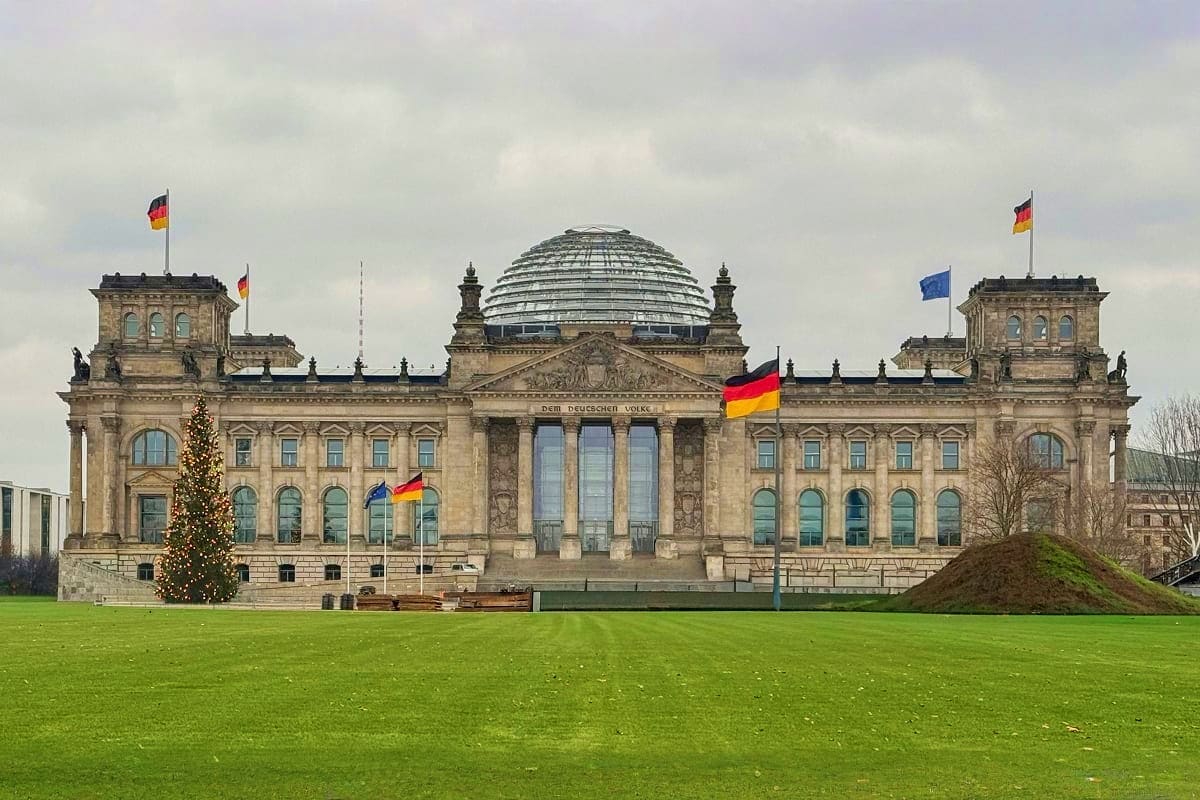 The Reichstag building in Berlin with German flags, glass dome, and manicured lawn in front under cloudy sky