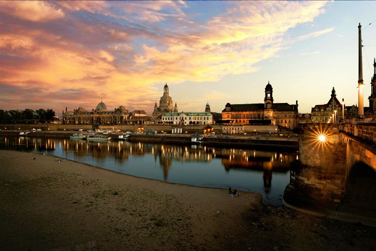 Dresden skyline at sunset with Elbe River, historic churches and buildings reflected in water, sandy beach in foreground
