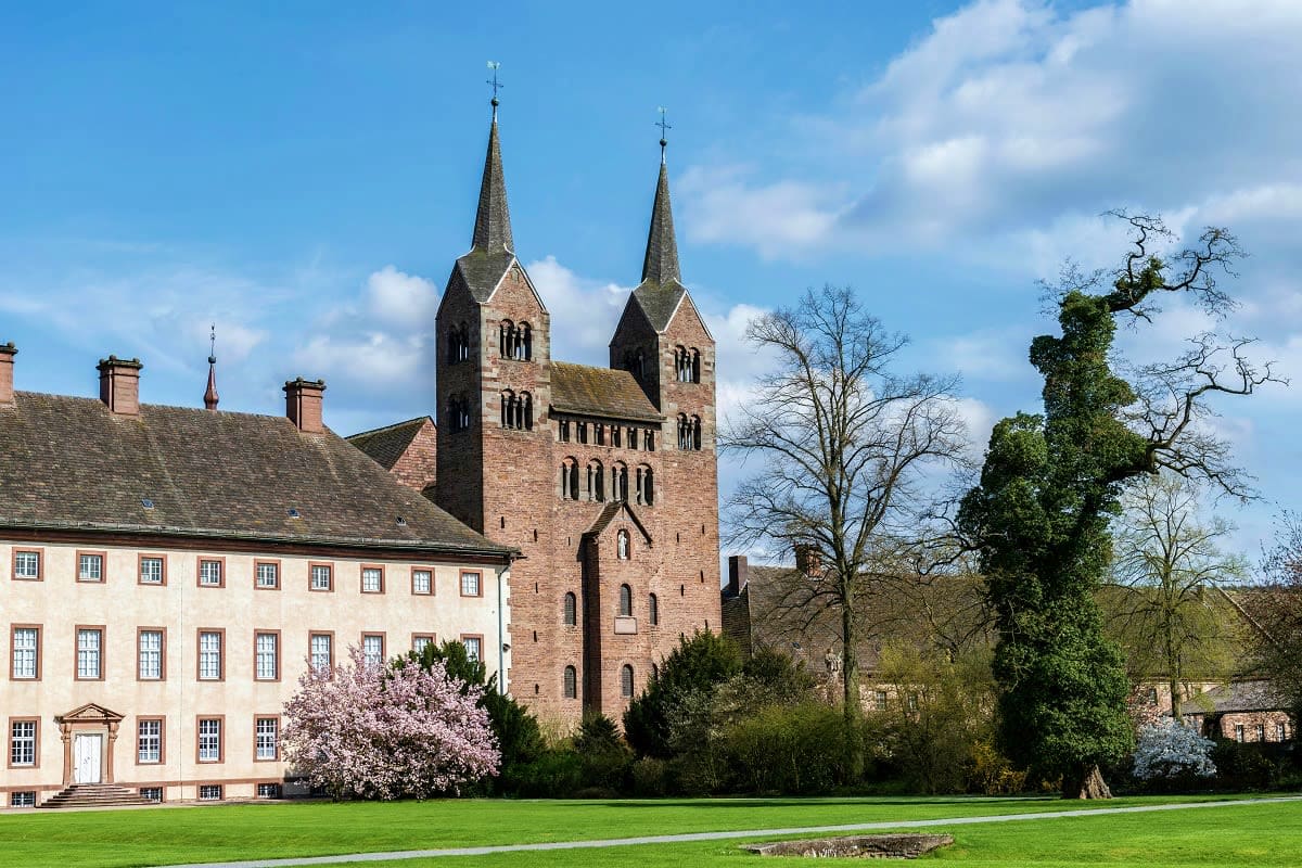 Historic German monastery with twin spires, adjacent cream-colored building, and spring cherry blossoms in landscaped grounds