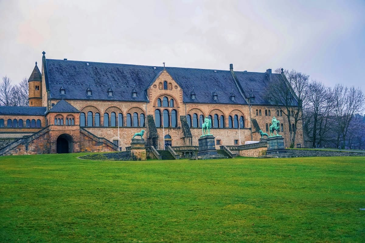 Historic red brick palace with slate roof and green copper statues on manicured lawn, leafless trees in background