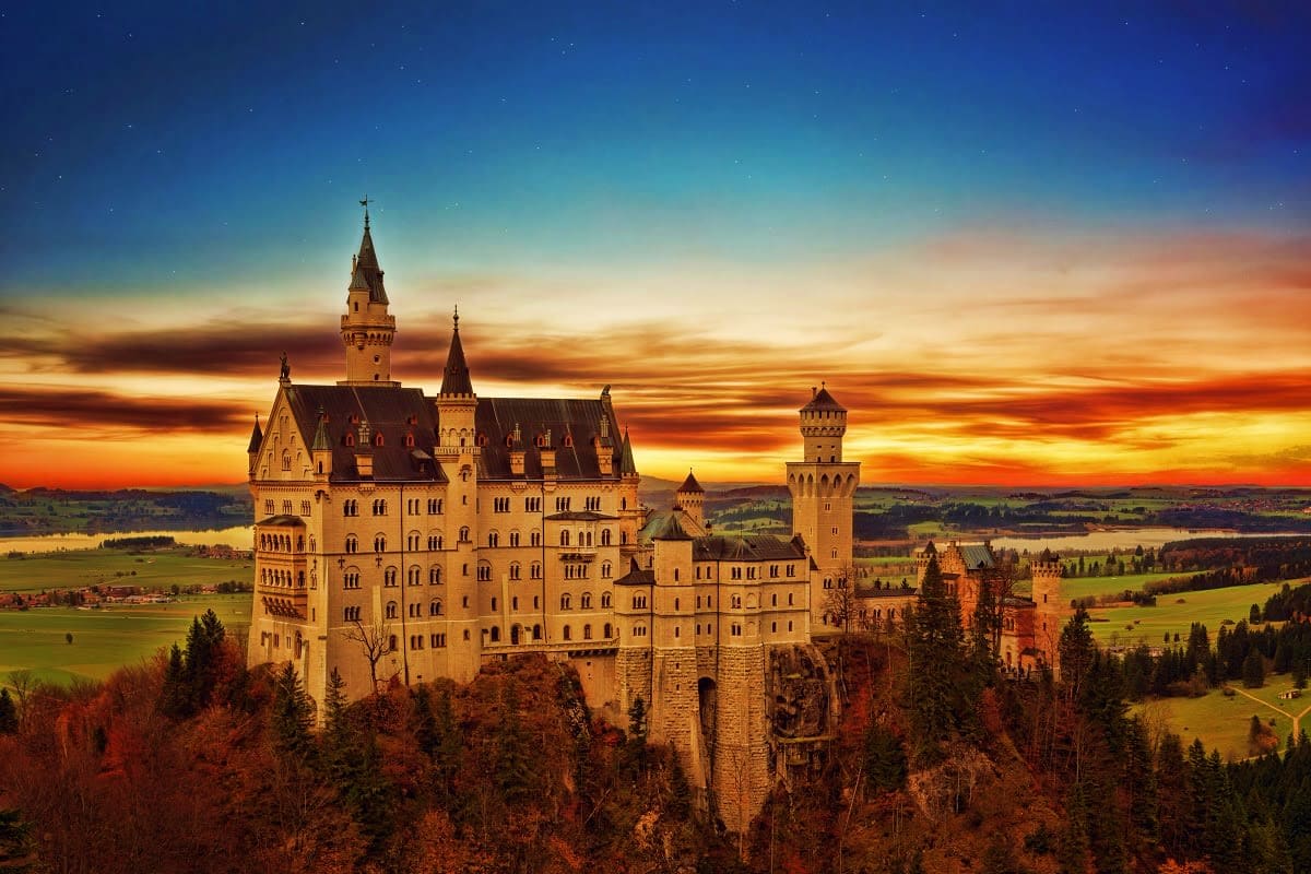 Neuschwanstein Castle perched on a clifftop at sunset with autumn forests and Bavarian countryside below under a starry sky