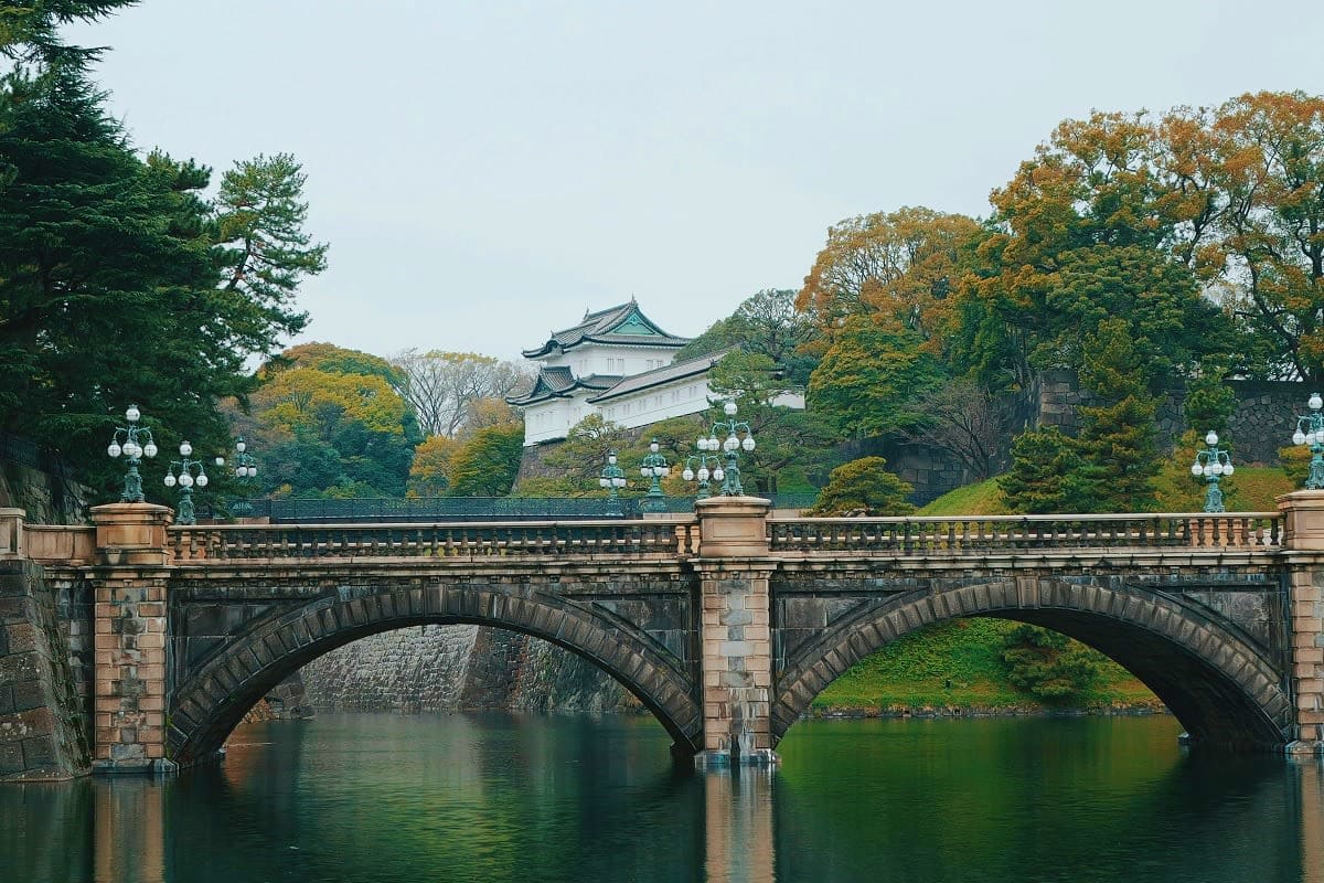 Stone bridge with multiple arches spanning a moat in front of a traditional Japanese castle surrounded by autumn foliage and 