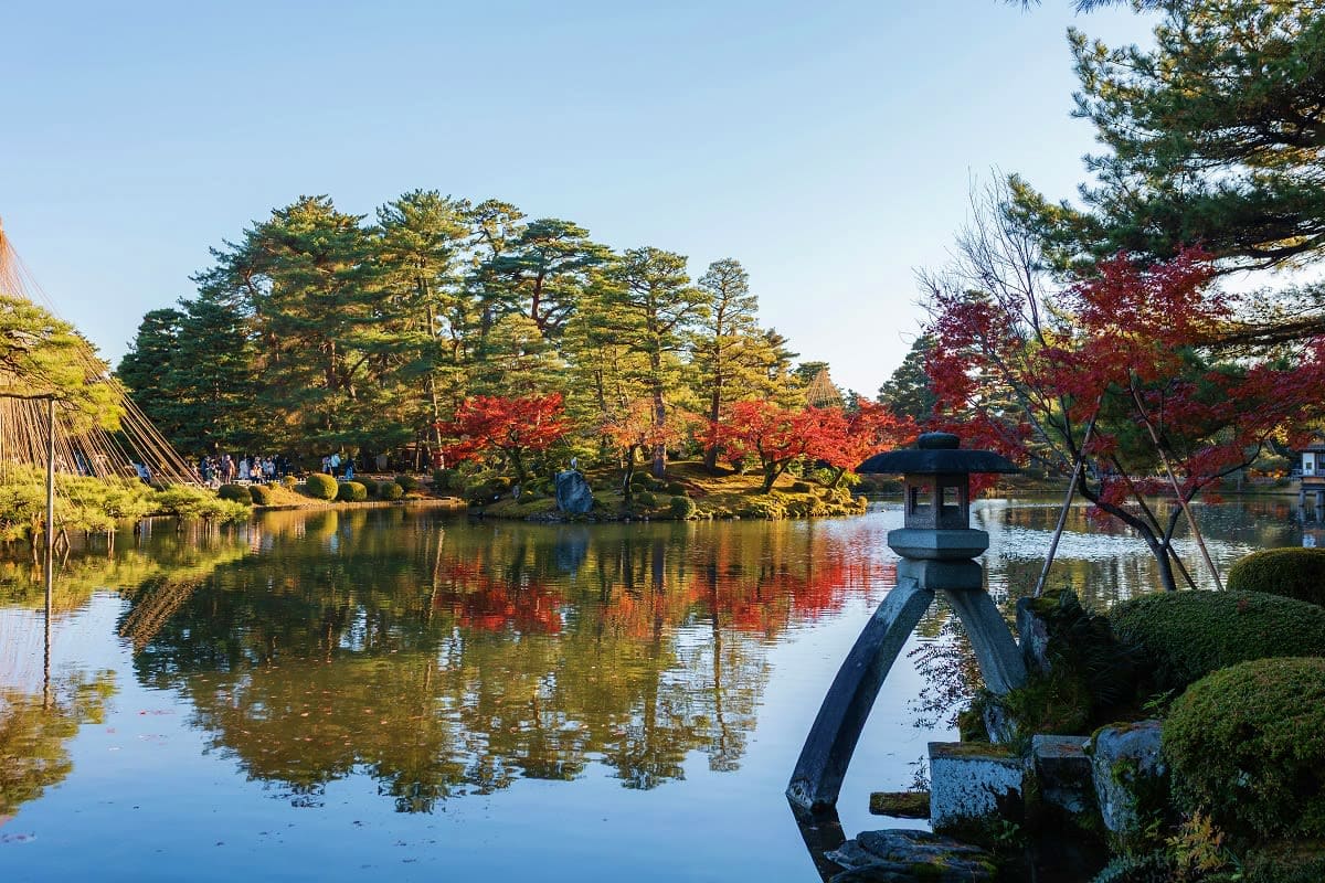 Japanese garden landscape with pond, stone lantern, autumn foliage in red and gold, traditional architecture and visitors exp