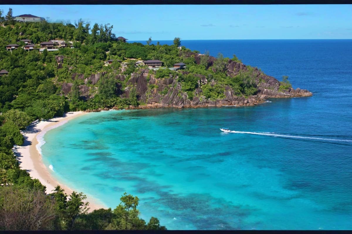 Aerial view of a tropical beach with turquoise waters, white sand, granite rock formations, lush vegetation, and luxury resor