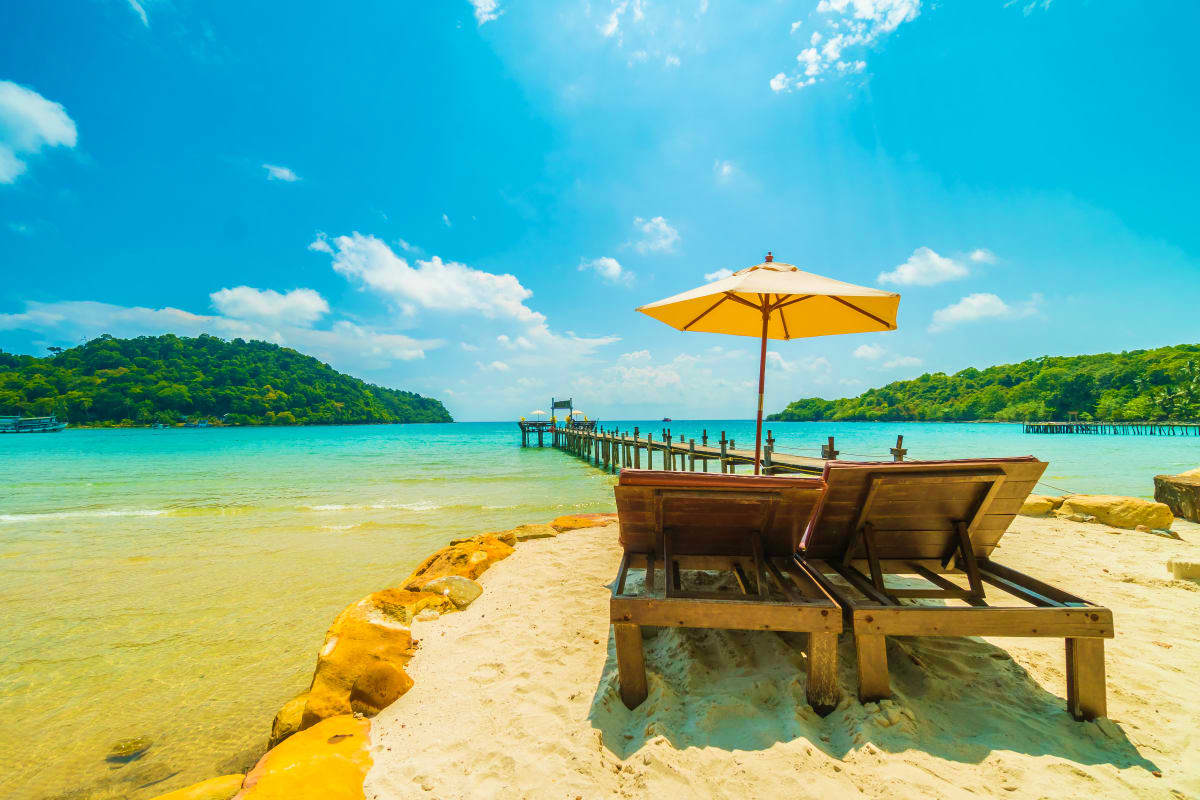 Wooden beach loungers with yellow umbrella on sandy shore overlooking turquoise lagoon, wooden pier, and forested islands in 