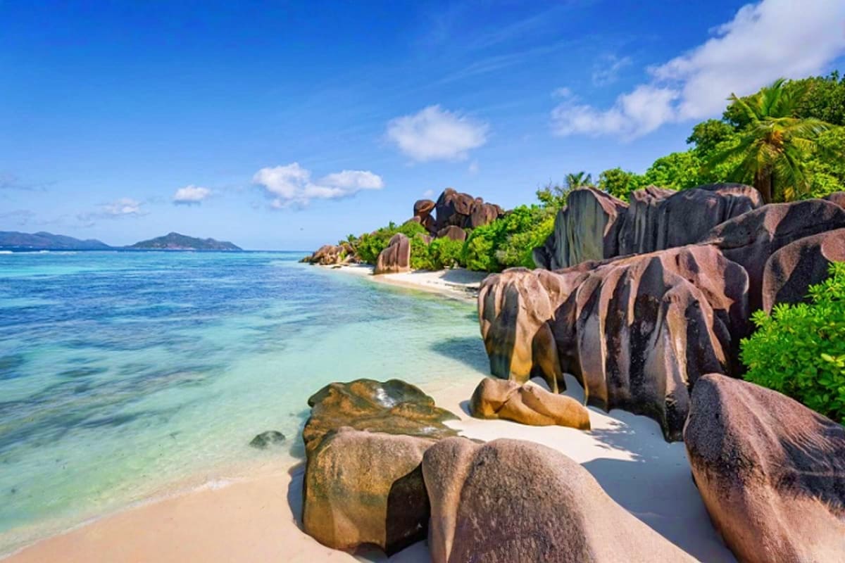 Tropical beach in Seychelles with granite boulders, turquoise water, white sand, and palm trees with distant islands under bl