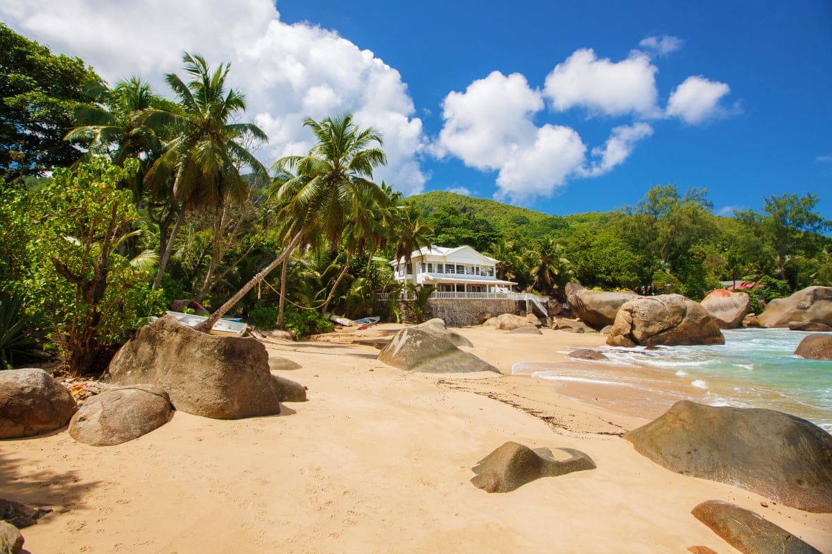 White beachfront villa on sandy beach with granite boulders, palm trees, and turquoise ocean waters in Seychelles