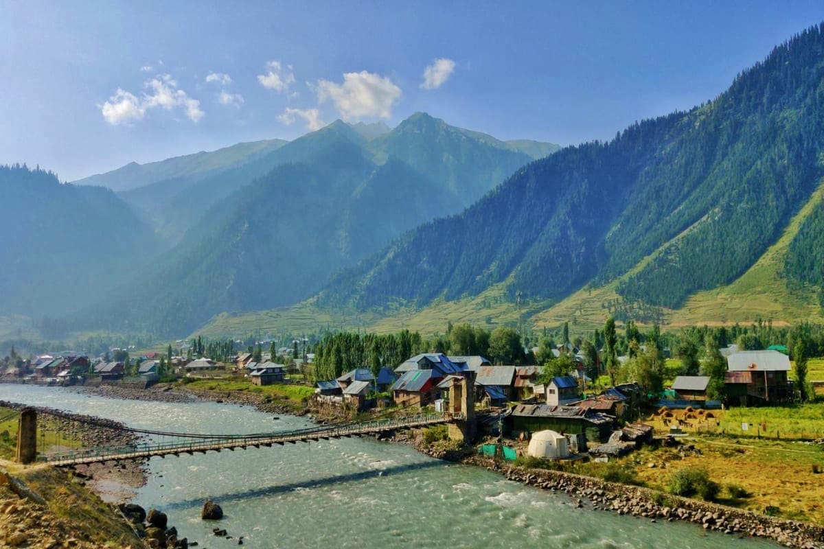 Mountain valley village in Kashmir with wooden houses, suspension bridge crossing turquoise river, forested peaks and snow-ca