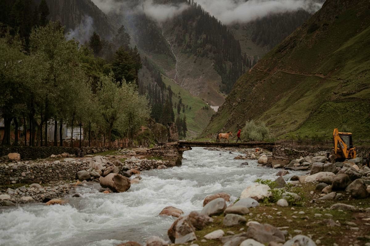 Rushing river flowing through mountainous Kashmir valley with pine trees, willow trees, traditional bridge, and person with h