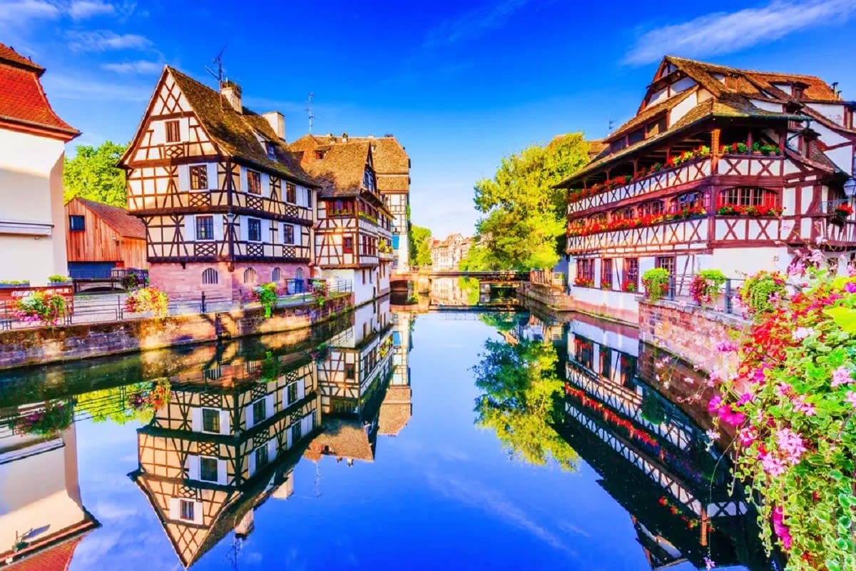 Historic half-timbered buildings reflected in calm canal water with blooming flowers and blue sky in Strasbourg, France