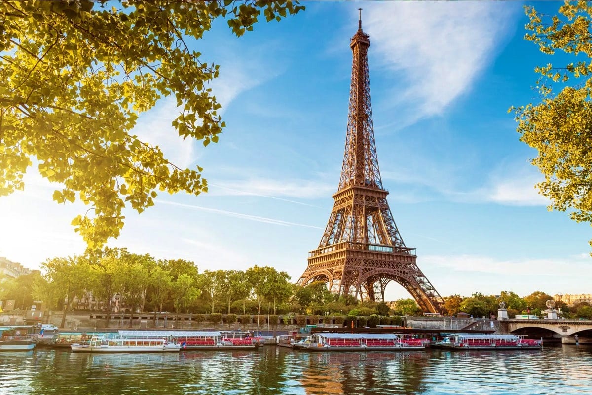 Eiffel Tower in Paris framed by golden autumn foliage with Seine River and tourist boats in foreground on blue sky day
