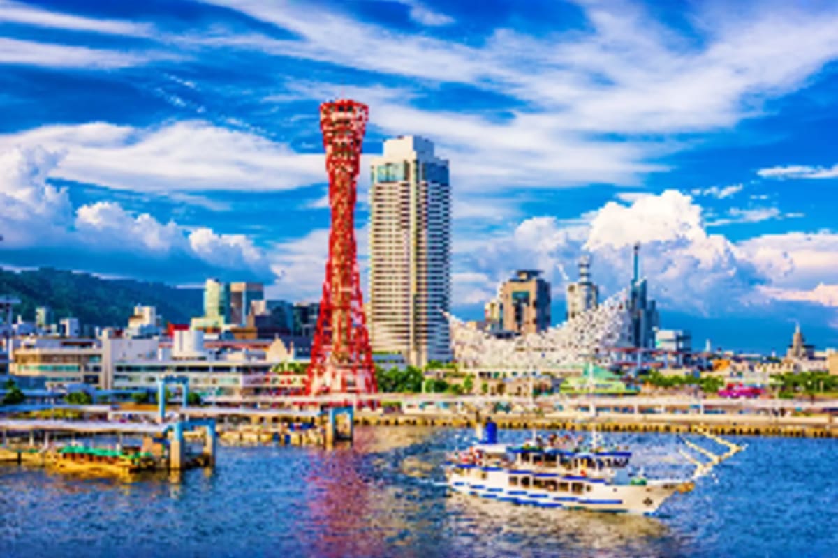 Kobe skyline with red Kobe Tower, modern skyscrapers, waterfront harbor with tour boats and blue ocean under cloudy sky