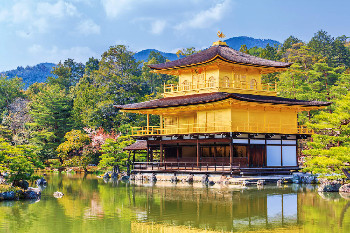 Golden Japanese temple with tiered roof reflected in calm pond, surrounded by lush green forest and mountains under blue sky