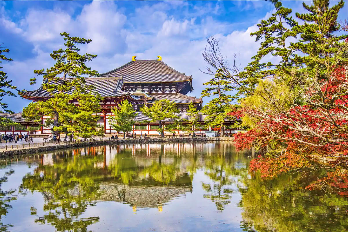 Traditional Japanese temple with curved roof and gold ornaments reflected in still pond, surrounded by autumn foliage and gre