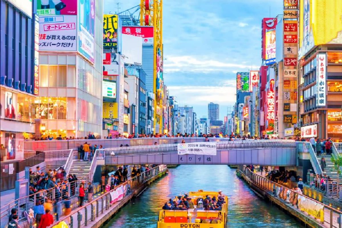 Dotonbori canal in Osaka with yellow sightseeing boat, crowded pedestrian bridge, and illuminated neon signs lining both side
