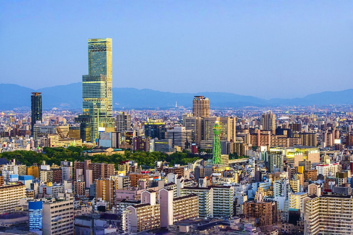 Osaka city skyline with modern skyscrapers, mountains in background, and dense urban landscape under clear blue sky