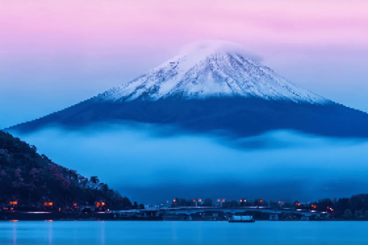 Snow-capped Mount Fuji at dawn with pink sky, mist-covered forested base, and lakeside town lights reflected in water below