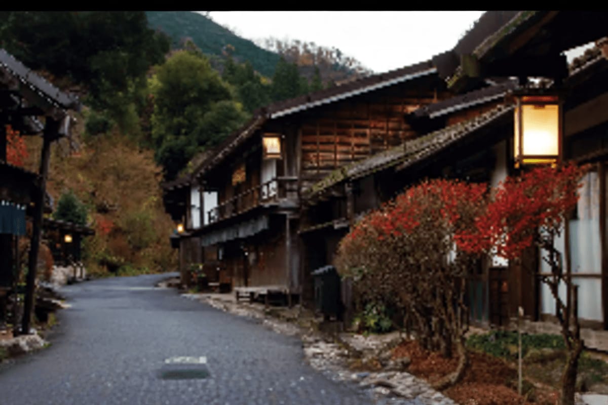 Traditional Japanese village street with wooden buildings, red autumn foliage, and mountains in background at dusk