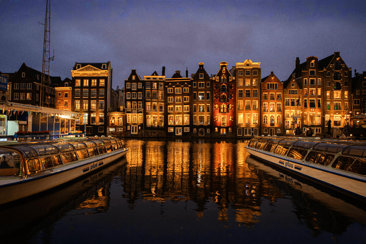 Amsterdam canal with illuminated historic merchant houses reflected in water at dusk, tour boats moored in foreground