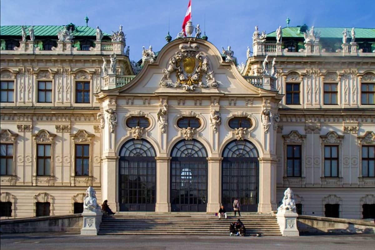 Ornate Baroque palace facade with green copper roofs, grand entrance archway, statues, and Canadian flag in Vienna