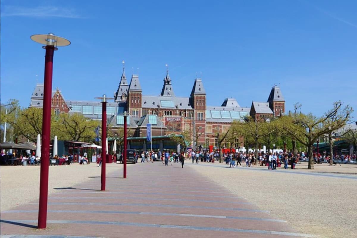 Amsterdam's Museumplein with Rijksmuseum building, red heating lamps, and crowds of visitors on a sunny day