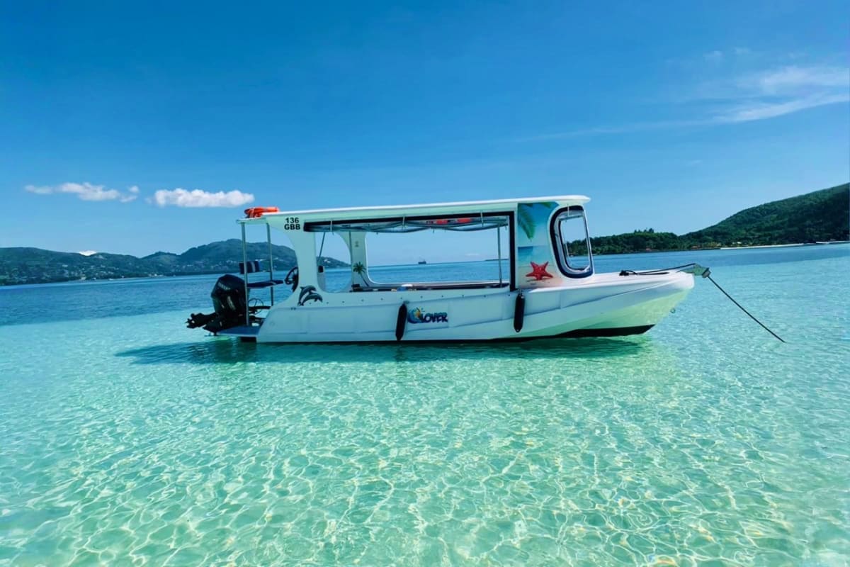 White glass-bottom boat moored in crystal-clear turquoise water with forested mountains in background, Seychelles