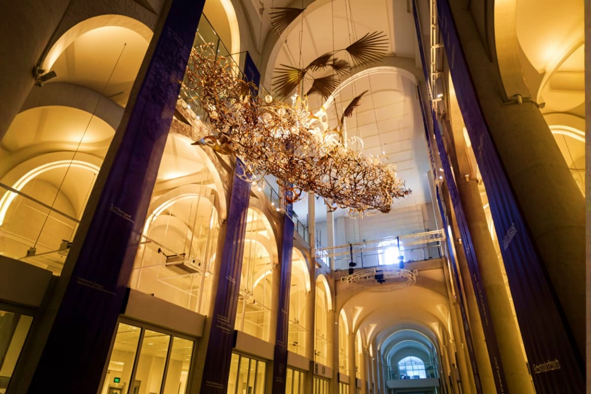 Explore Manila And Cebu at Your Own Pace Modern gallery hallway with soaring arched ceilings, warm lighting, dark columns, and suspended bronze sculptural installatio
