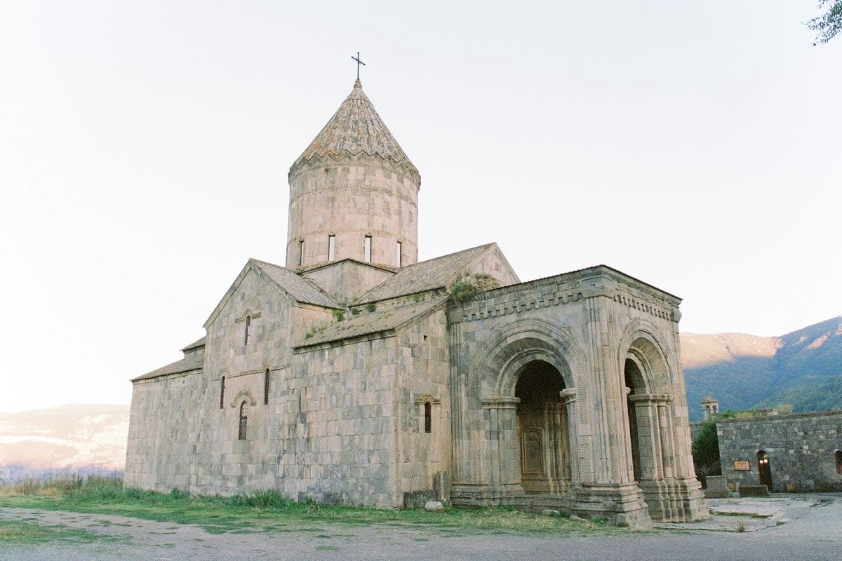Ancient Armenian church with conical dome and cross, stone arches, mountainous landscape in background