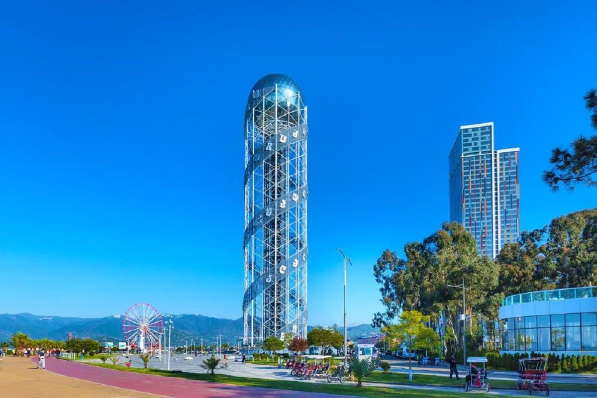 Modern cylindrical tower with geometric lattice design beside residential buildings in Tbilisi waterfront park with Ferris wh