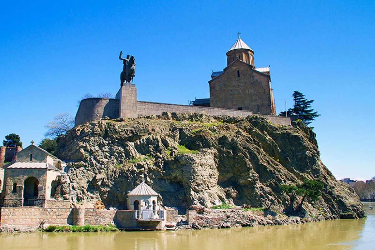 Metekhi Church and equestrian statue perched on rocky cliff overlooking Mtkvari River in Tbilisi, Georgia under clear blue sk