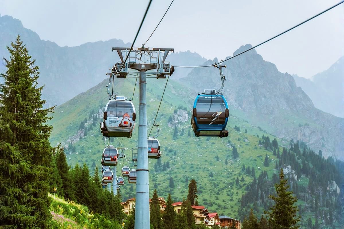 Cable car gondolas suspended above forested mountains and alpine valley with scattered buildings in Kazakhstan
