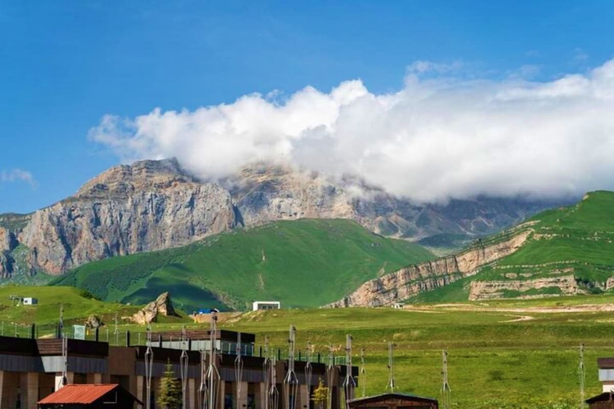 Mountain landscape in Azerbaijan with green meadows, traditional wooden fence, and snow-capped Shahdag peak partially obscure
