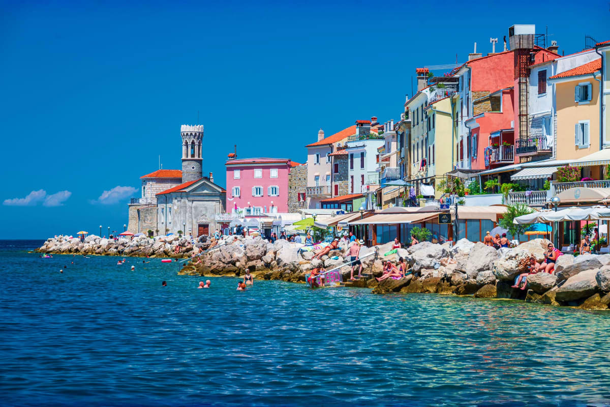 Colorful waterfront buildings and swimmers on rocky beaches along the Adriatic coast with historic church tower and clear blu