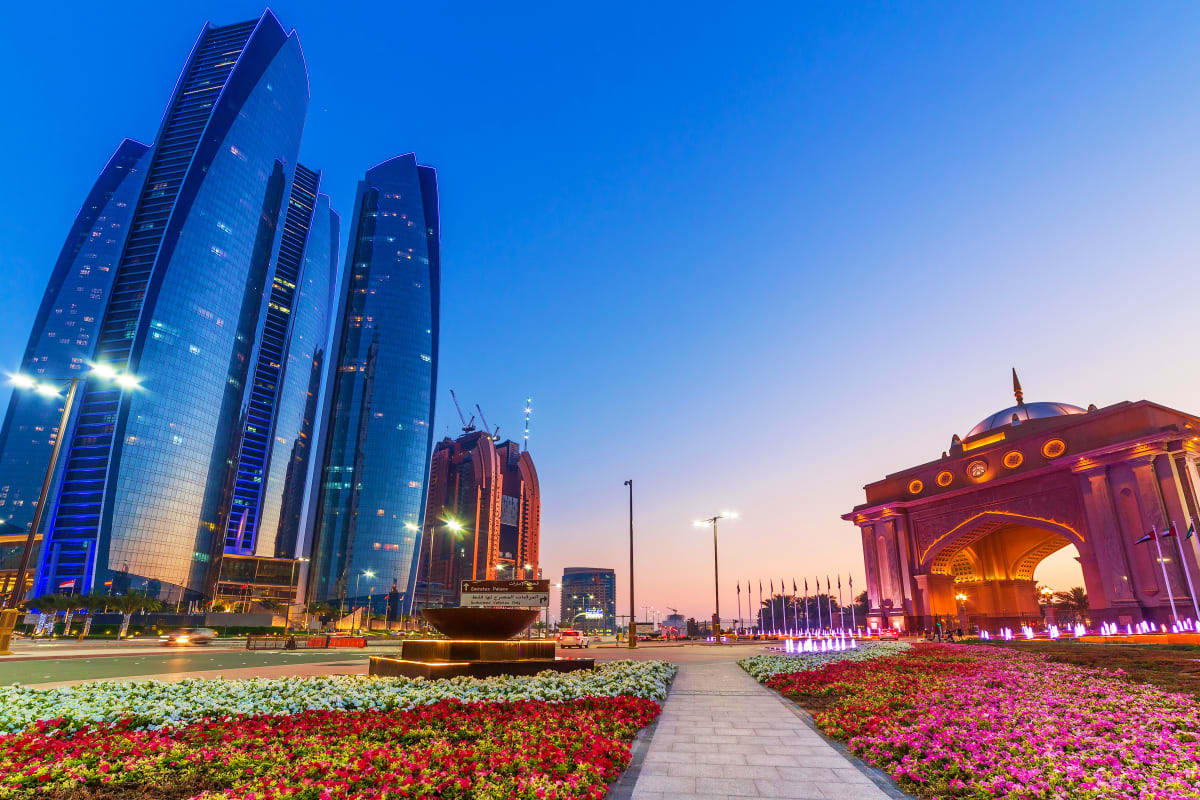 Abu Dhabi Stay And Play Luxury Package Modern skyline of Abu Dhabi at dusk with illuminated skyscrapers, colorful flower gardens, and the Sheikh Zayed Grand Mosque
