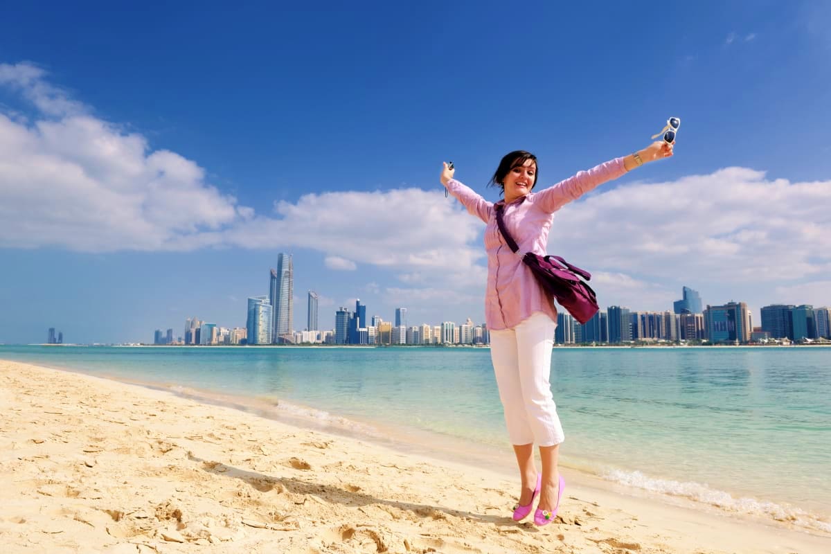 Abu Dhabi Stay And Play Luxury Package Woman with arms raised holding binoculars on Abu Dhabi beach with city skyline in background