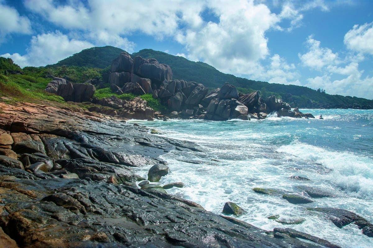 Amazing Seychelles - 05N & 06D Rocky granite boulders and turquoise ocean waves at a tropical Seychelles beach with forested mountain backdrop