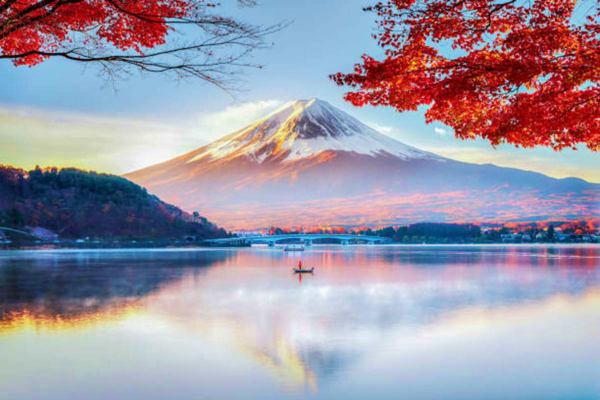 Mount Fuji reflected in Lake Kawaguchi during autumn with red maple leaves framing the snow-capped volcano and a lone fisherm