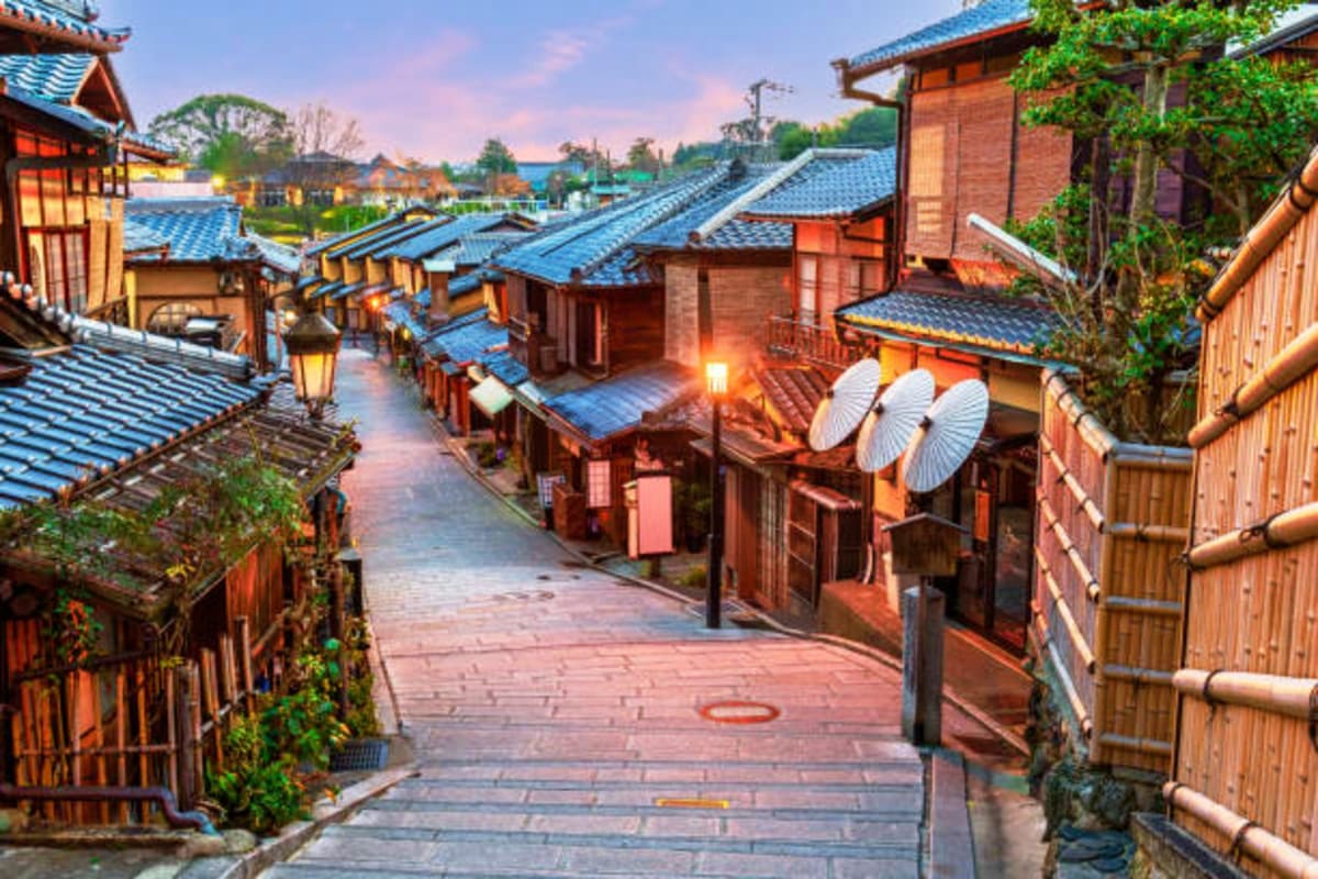 Traditional Japanese village street at dusk with wooden buildings, blue-tiled roofs, lanterns, and white umbrellas