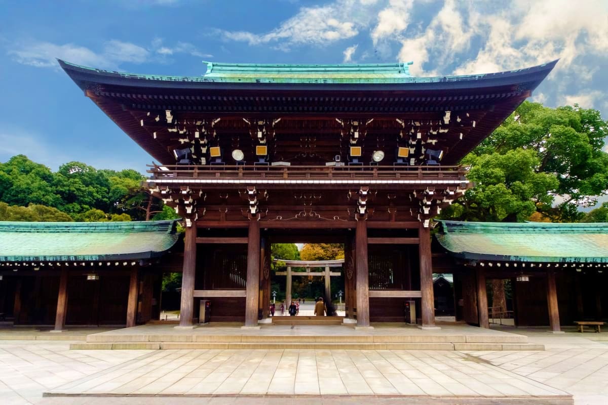Traditional Japanese temple with dark wooden architecture, curved green roof, and stone courtyard surrounded by trees under b