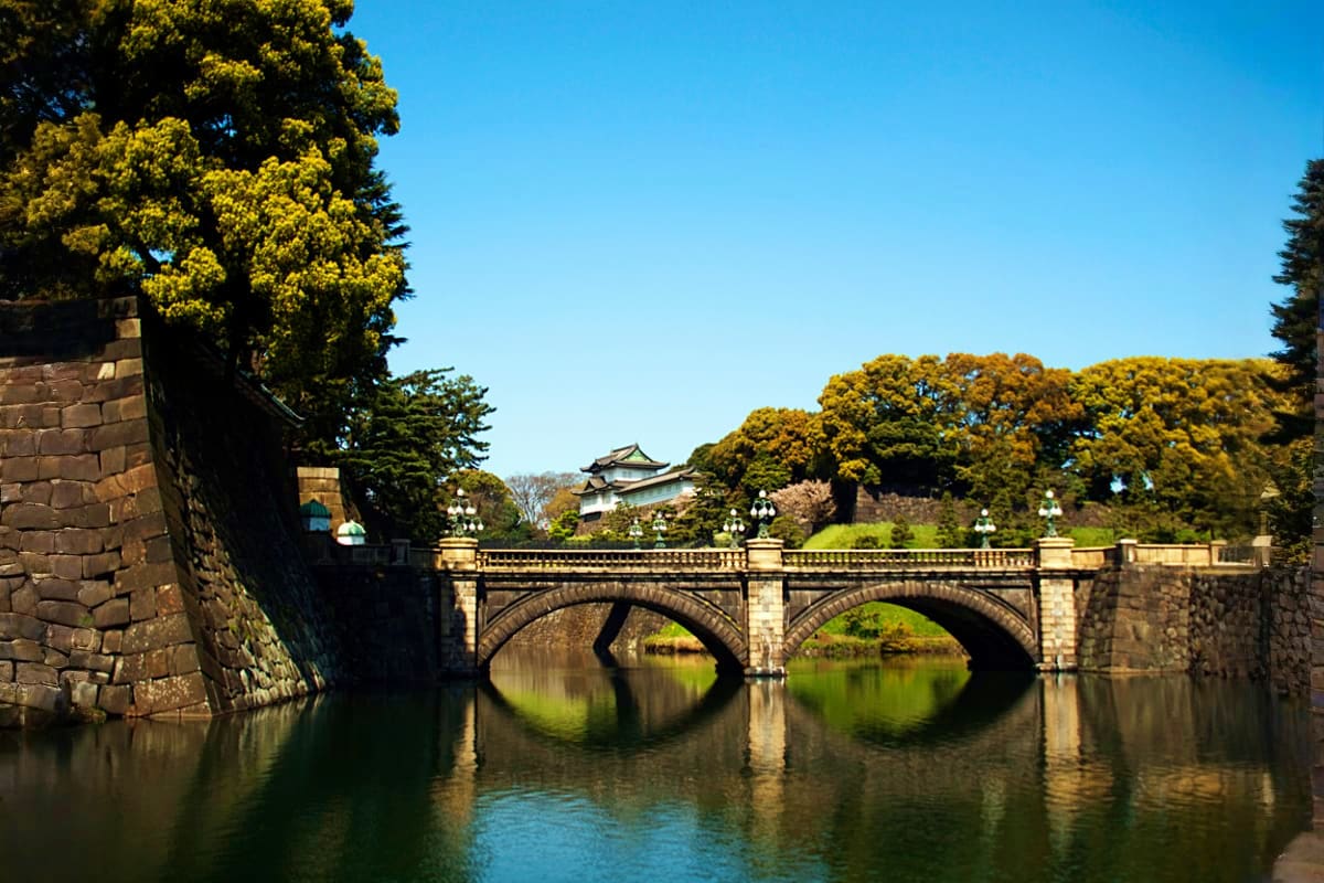 Stone bridge with two arches spanning calm water, traditional Japanese castle tower visible beyond lush green trees under cle
