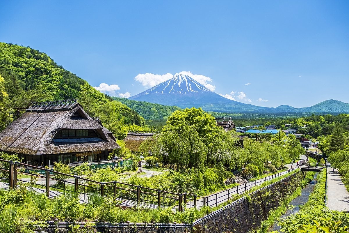 Traditional Japanese village with thatched-roof house overlooking Mount Fuji and lush green valleys under clear blue sky