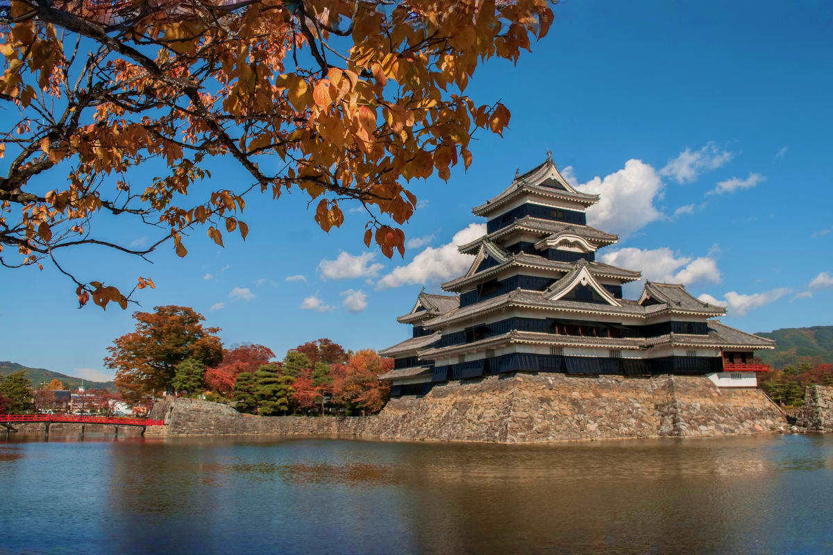 Matsumoto Castle with autumn foliage framing the view across the moat, clear blue sky, and fall colors in surrounding landsca