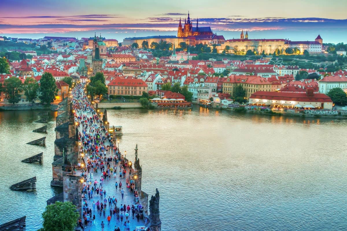 Prague cityscape at sunset with Charles Bridge crowded with tourists, Vltava River, and illuminated Prague Castle in backgrou