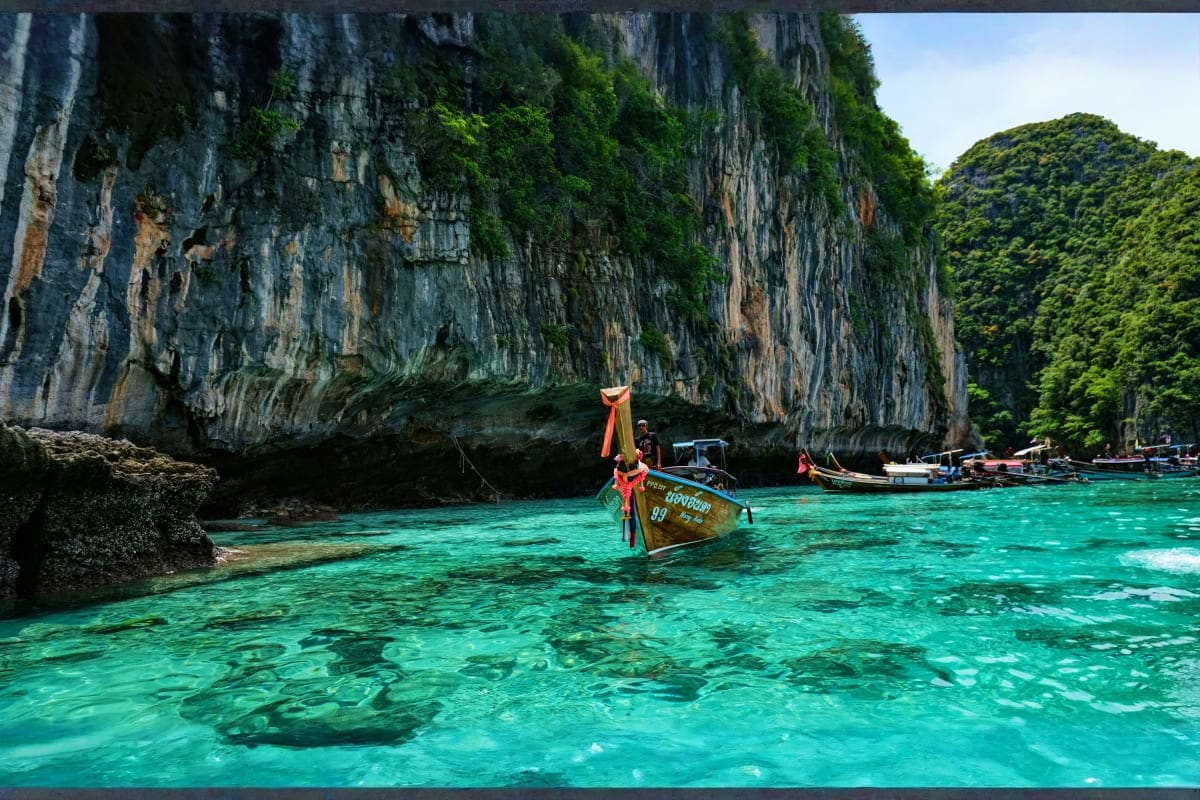 Traditional wooden long-tail boat in turquoise water beneath towering limestone cliffs in Thailand's Phi Phi Islands