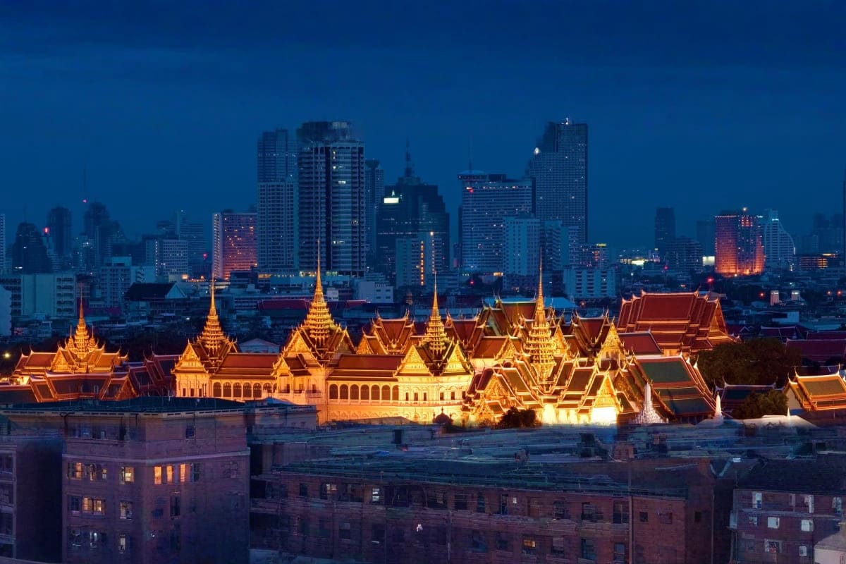 Grand Palace illuminated at dusk with Bangkok skyline and modern skyscrapers in background at twilight