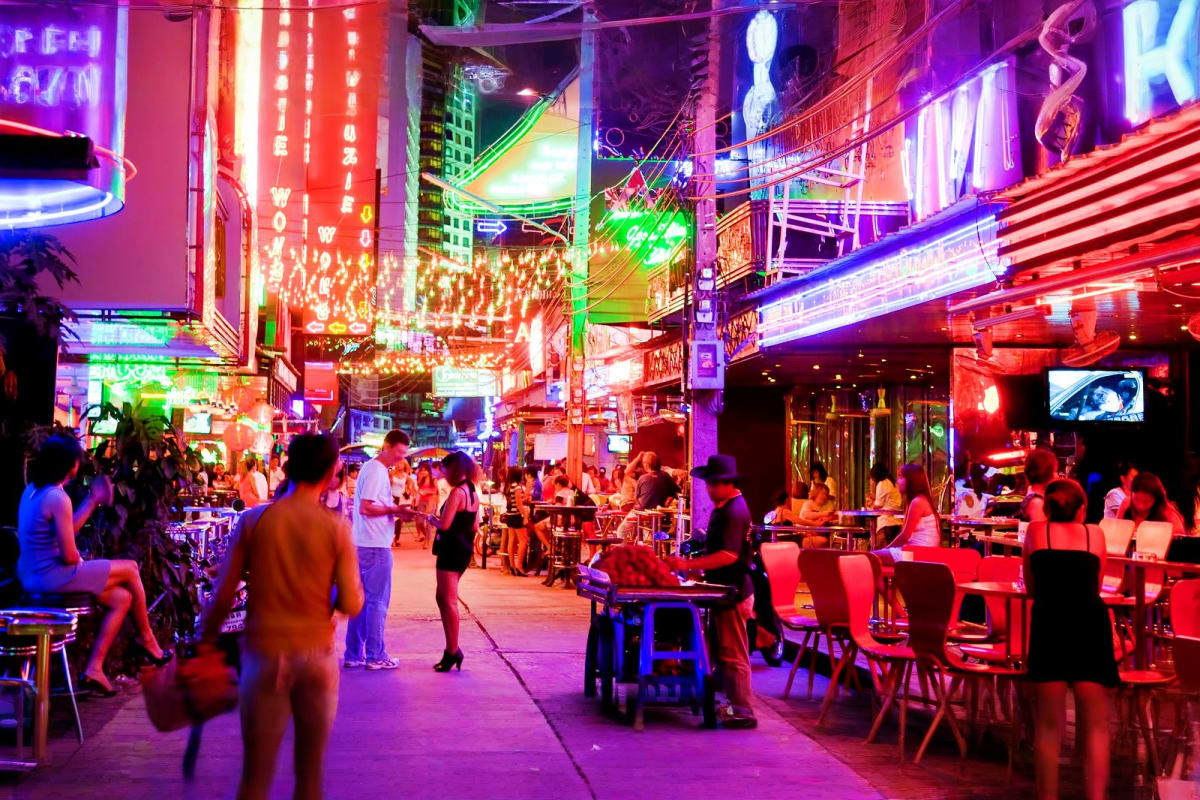 Vibrant nighttime street scene with neon signs in red, purple, and green lighting, crowds of people walking and sitting at ou