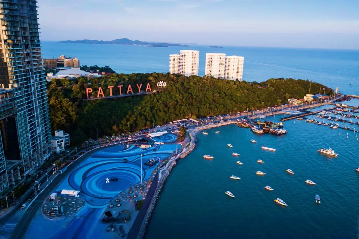 Aerial view of Pattaya City beachfront with blue water park, marina boats, high-rise hotels, and forested hillside with "Patt